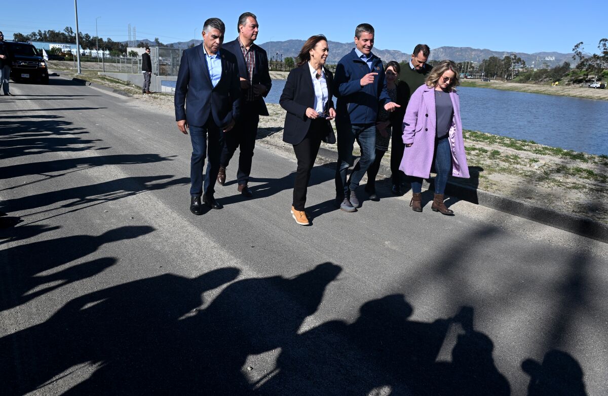 A woman in a pantsuit walks with five other people on a paved walkway alongside a body of water.