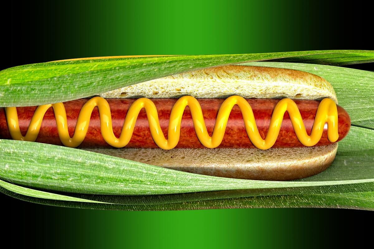 A hot dog in a bun, squiggled with mustard, nestled into green leaves