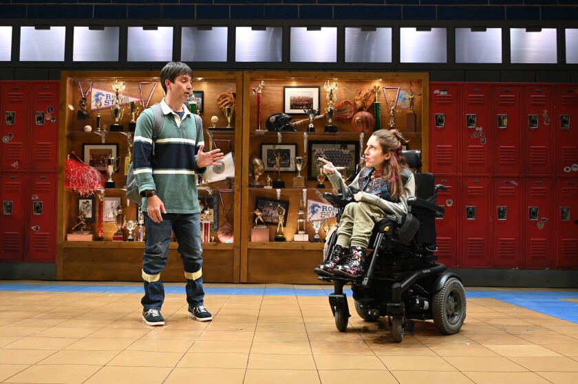 A boy stands talking to a girl in a wheelchair in a high school hallway