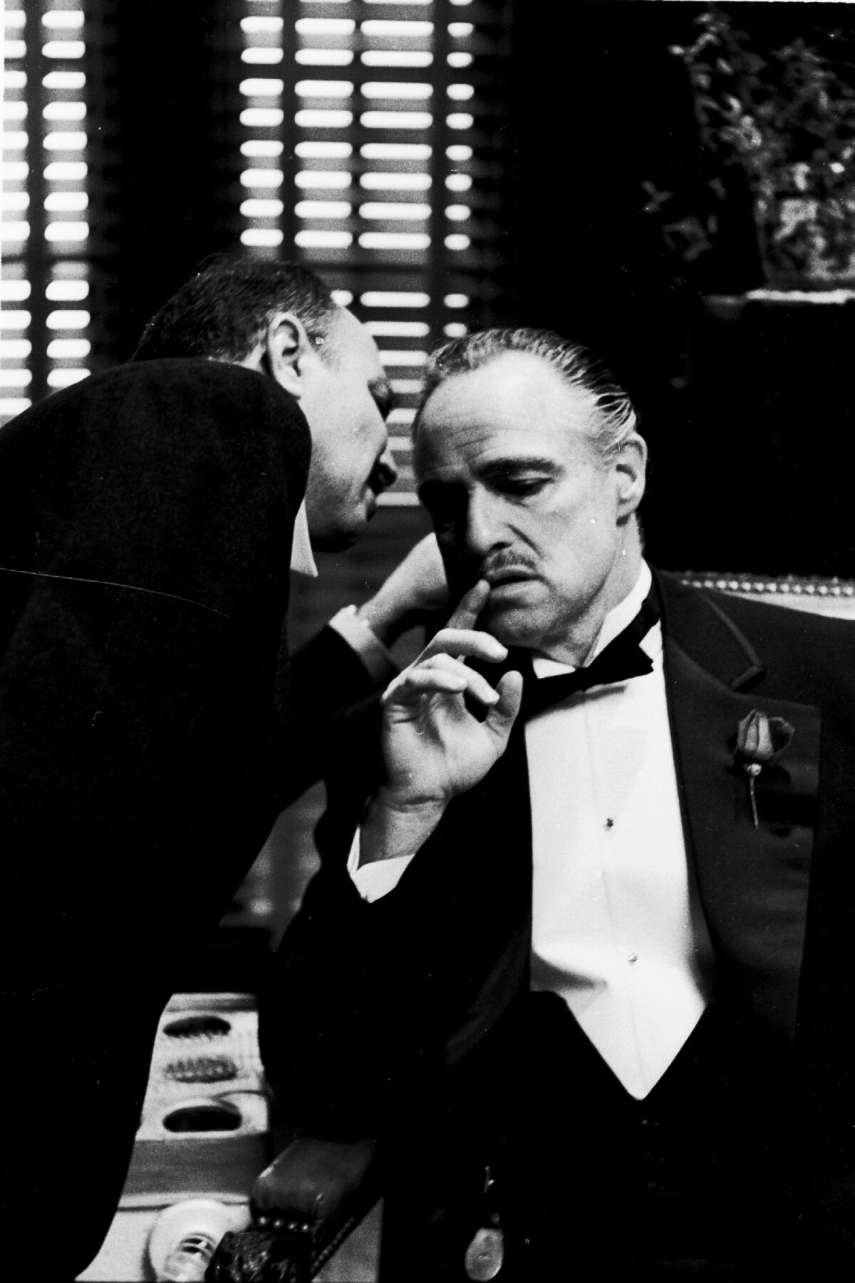 A man whispers into the ear of another man who is wearing a tuxedo.
