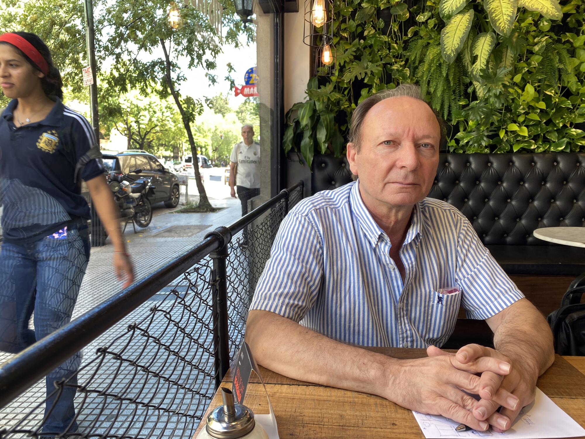 A man in a striped short-sleeve shirt, seated at a table with hands clasped