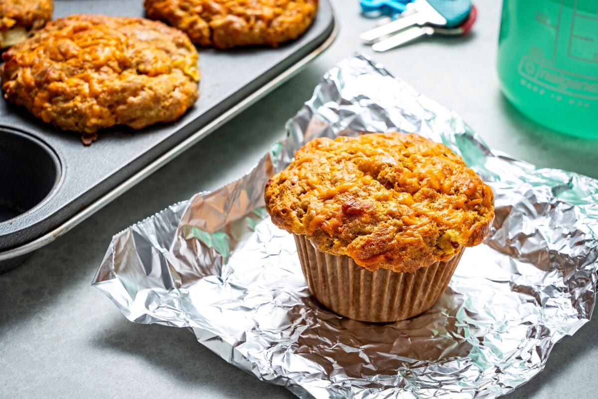 A freshly baked apple cheddar muffin sits on a piece of foil, with the muffin tin containing more next to it.