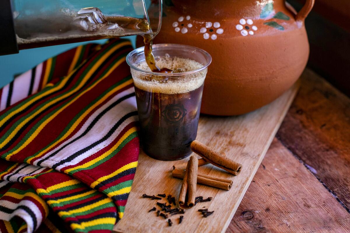 An iced coffee drink sits next to a colorful blanket, a ceramic pot and cinnamon sticks