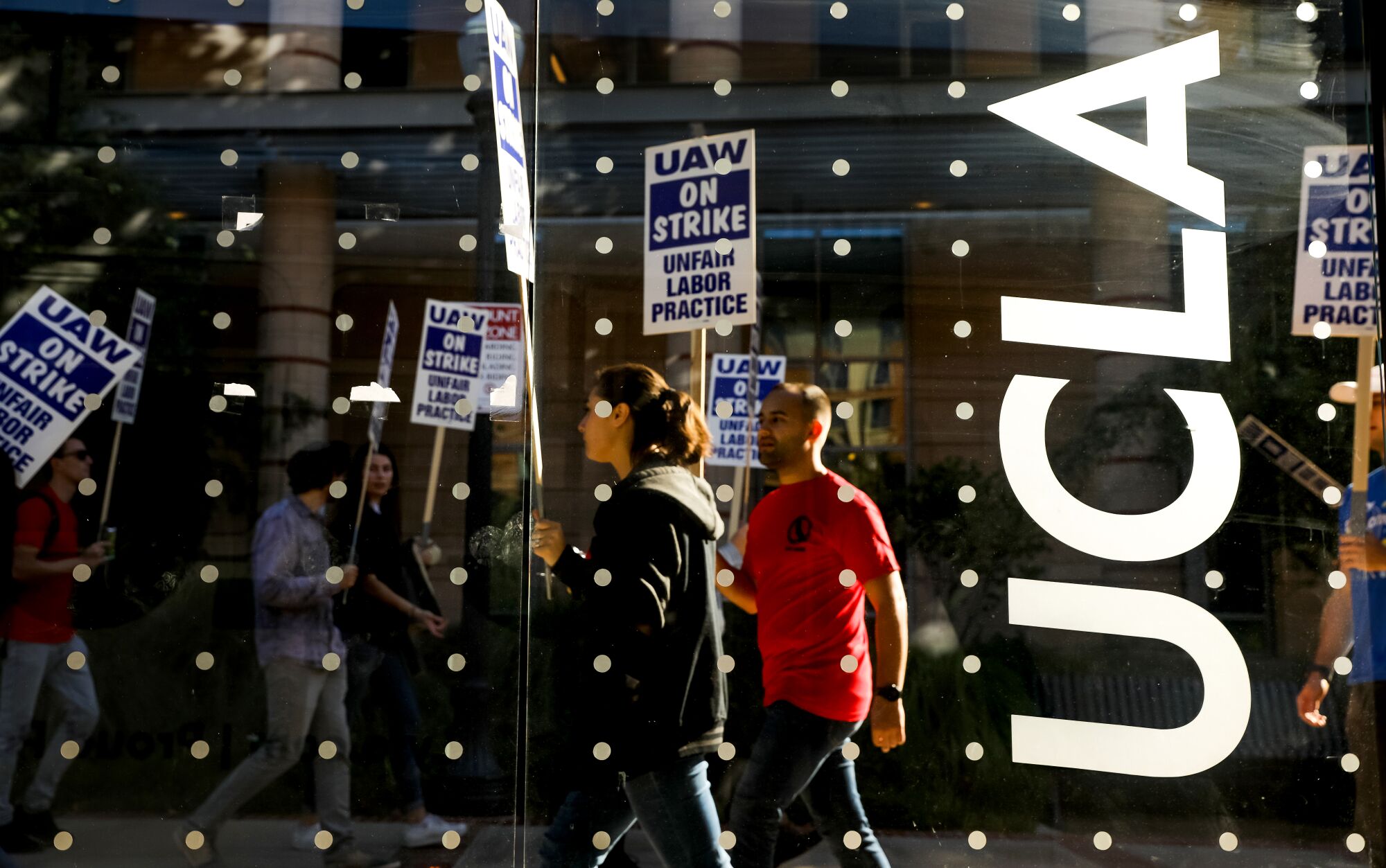 Demonstrators picket at UCLA near a window with dots and the UCLA logo on it.
