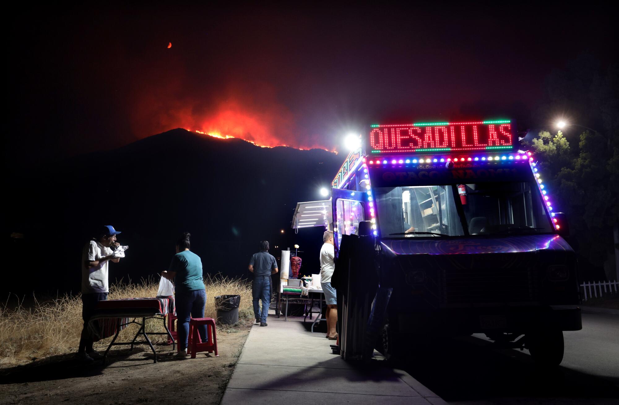 People eat by a taco truck as a wildfire illuminates a ridge in the distance.