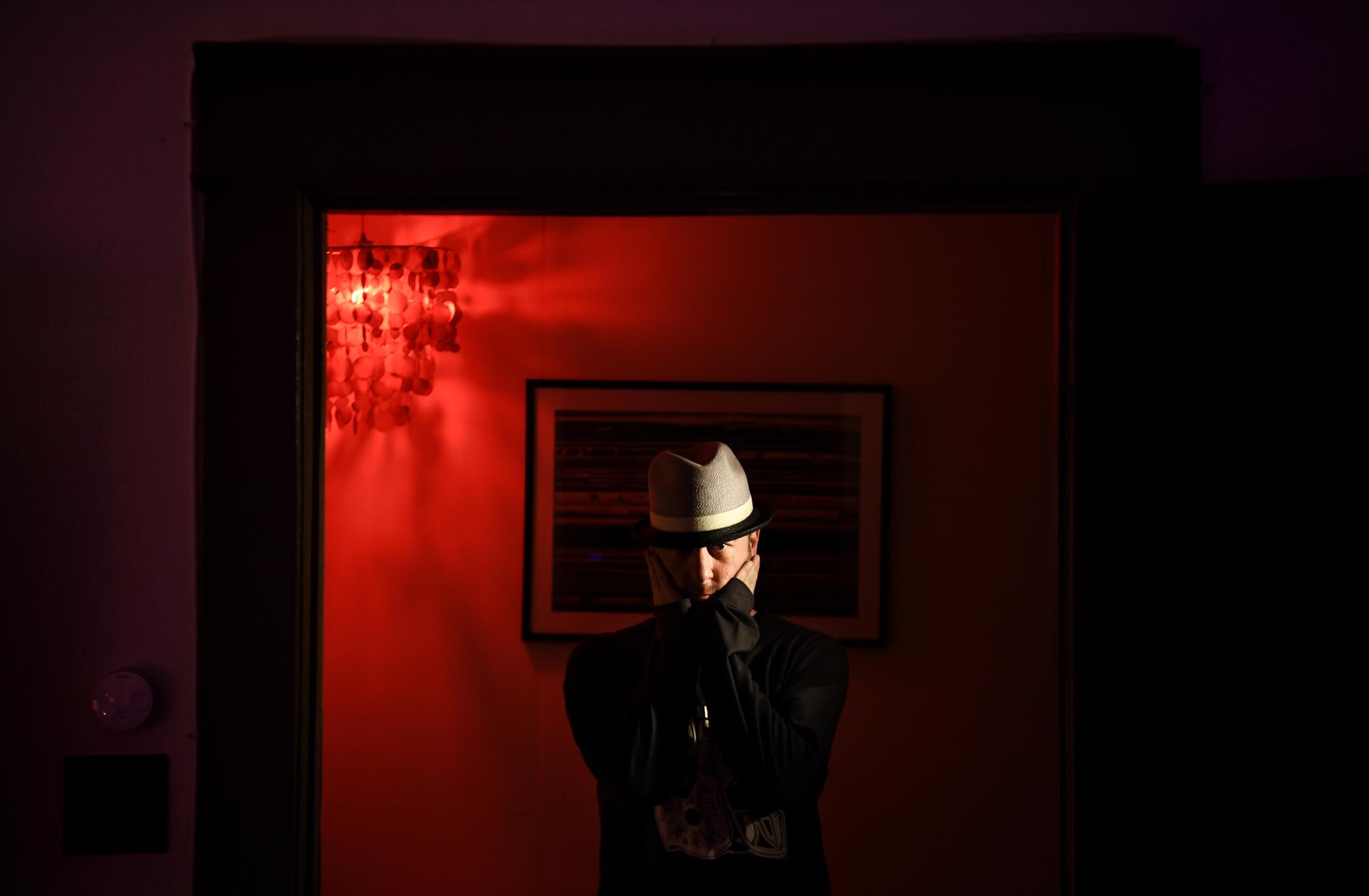 A man poses for a portrait with his hands on his face in a dark, red-lighted studio