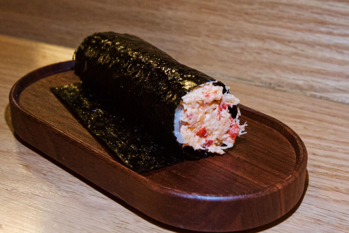 A single hand roll, stuffed with shredded lobster, sitting on an oval wood plate