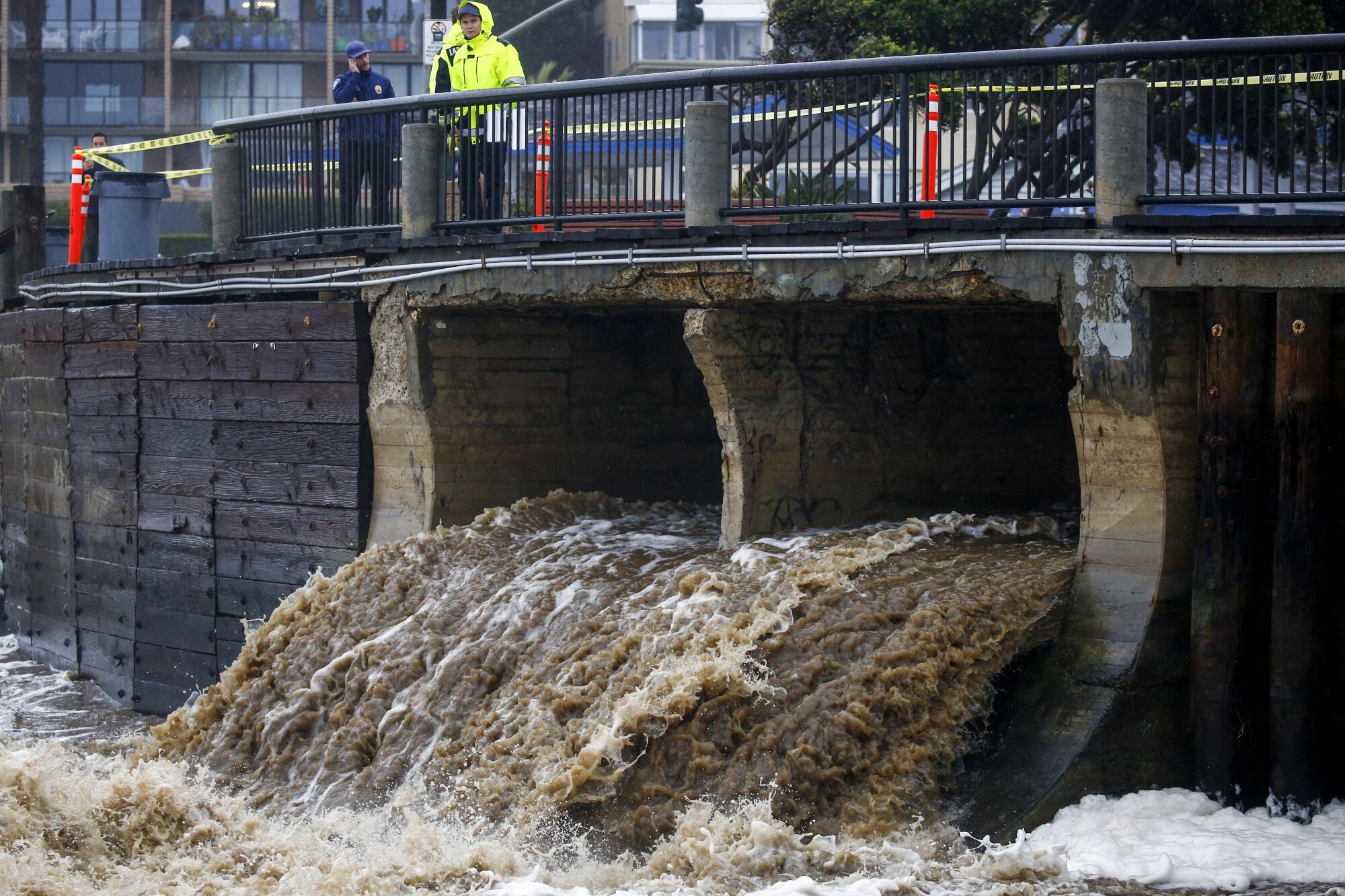 Stormwater falls into the ocean from a drainage canal as passersby watch.