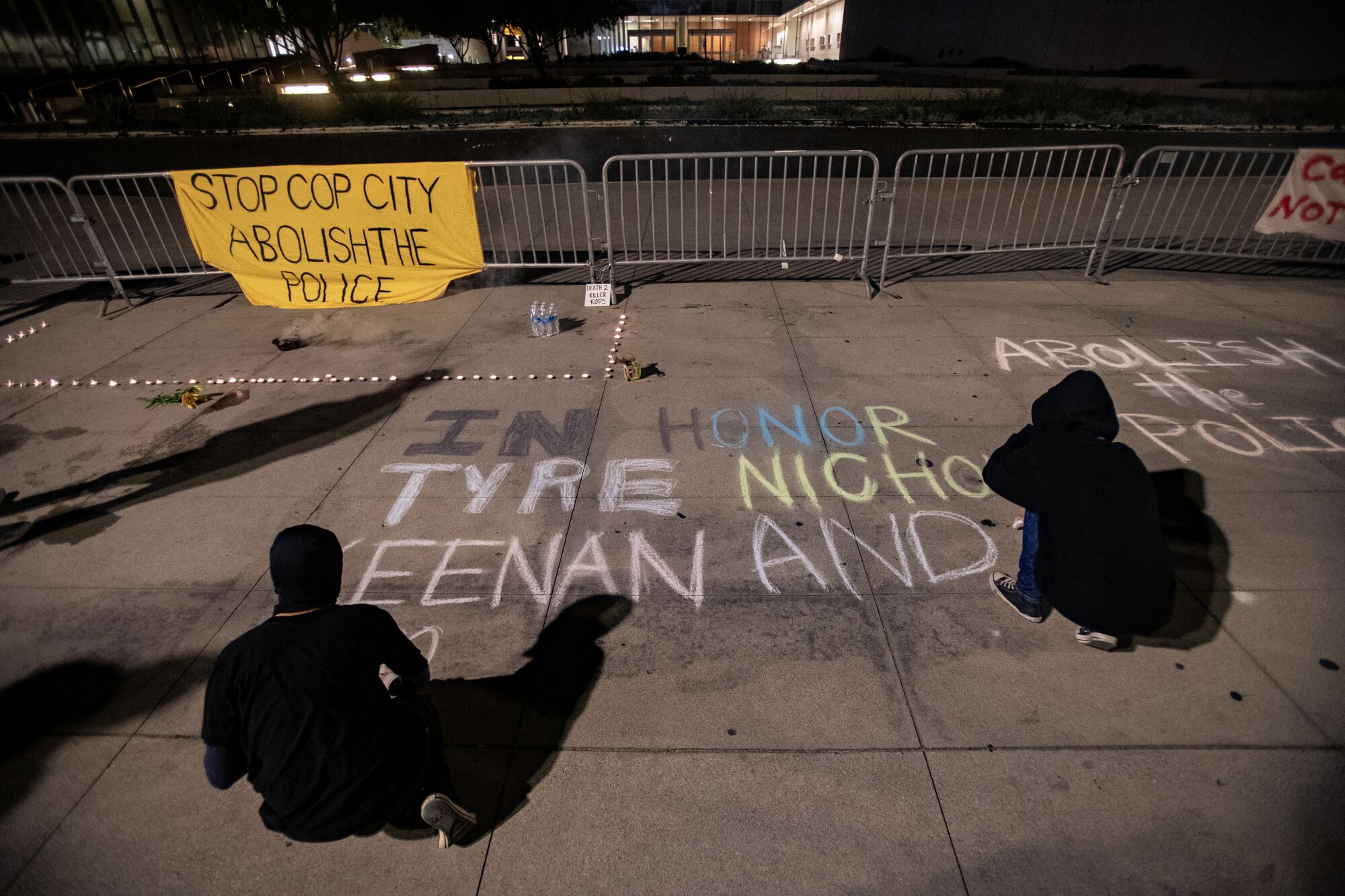Two people, dressed in black sweatshirts, write "In honor. Tyre Nichols. Keenan Anderson" on the sidewalk