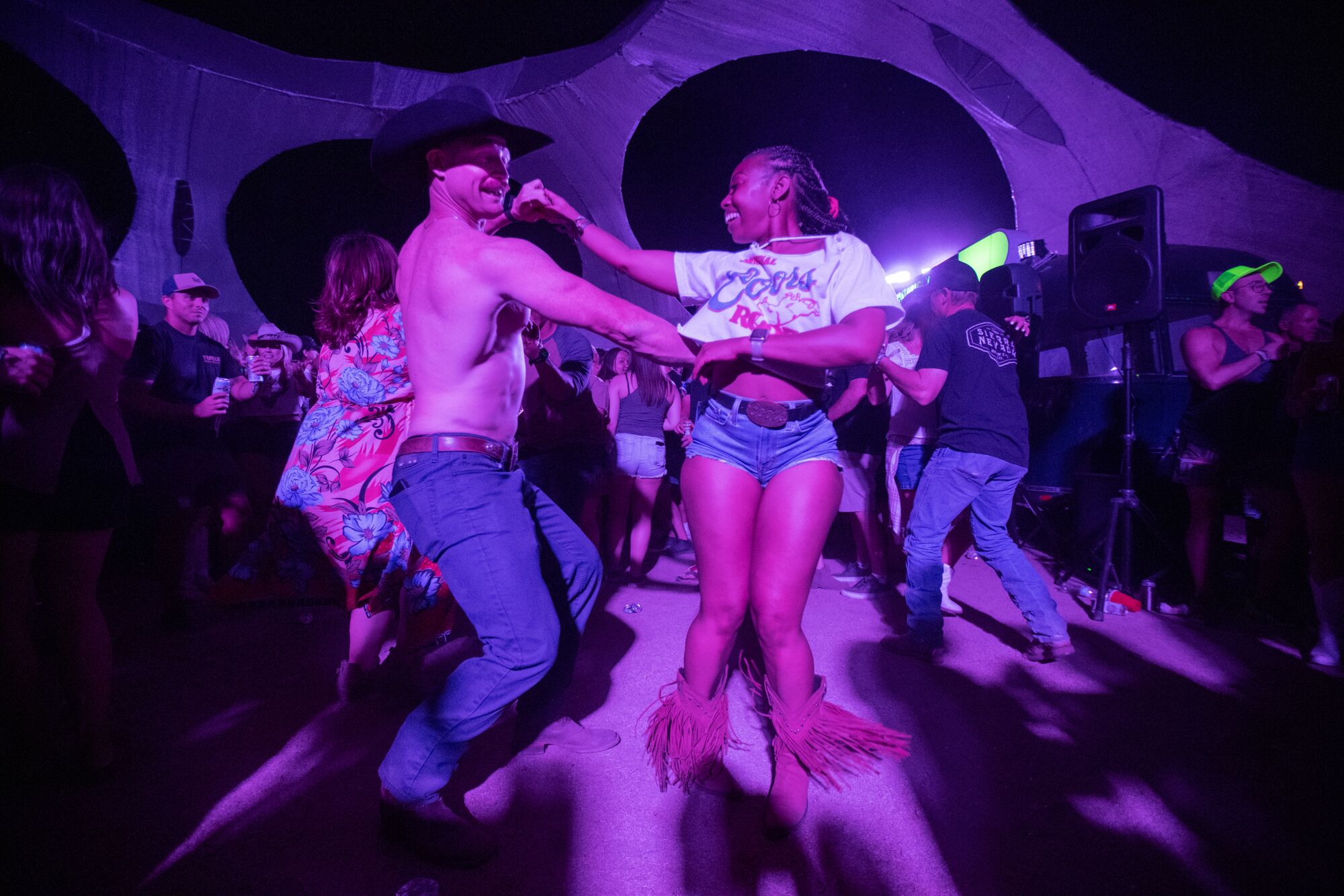 Illuminated by blue and magenta, a couple is holding hands dancing among others. 