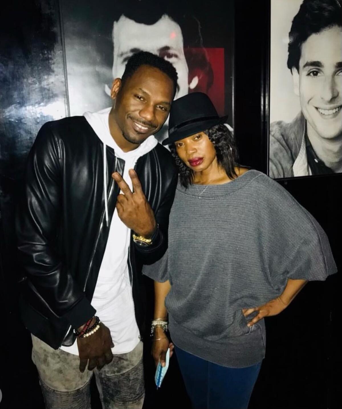 A man and a woman pose together inside a club with pictures of comedians on a dark wall in the background.