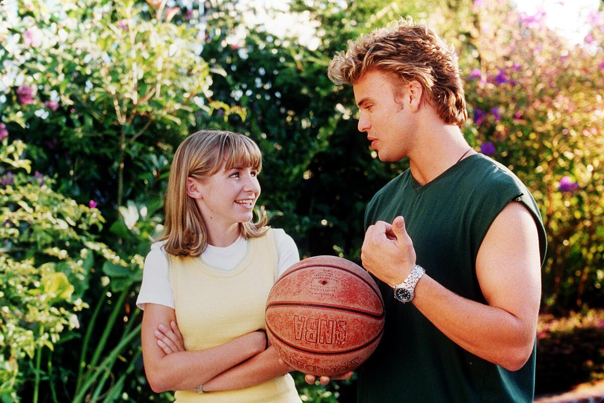 Una niña con una camiseta amarilla parada junto a un adolescente con una camiseta verde sin mangas sosteniendo una pelota de baloncesto.