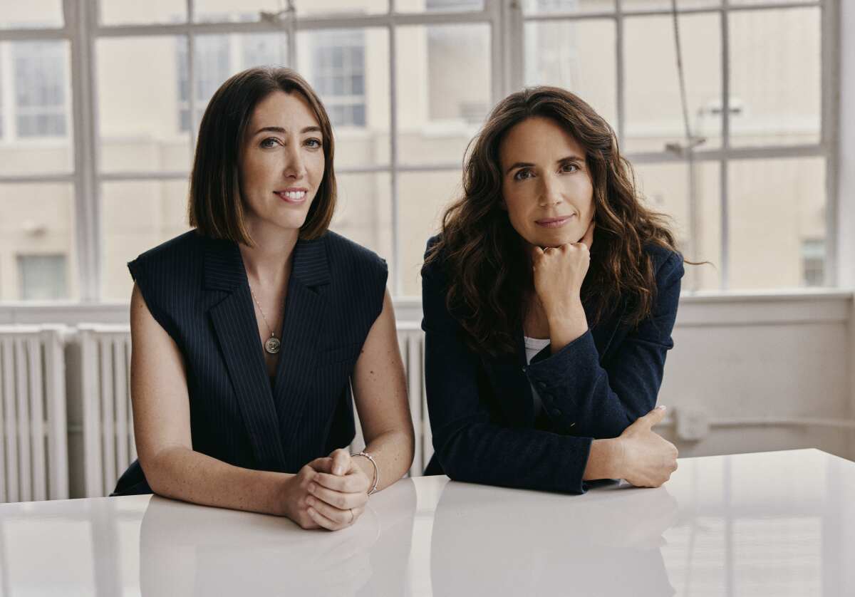 Two women in dark outfits sitting at a white table.