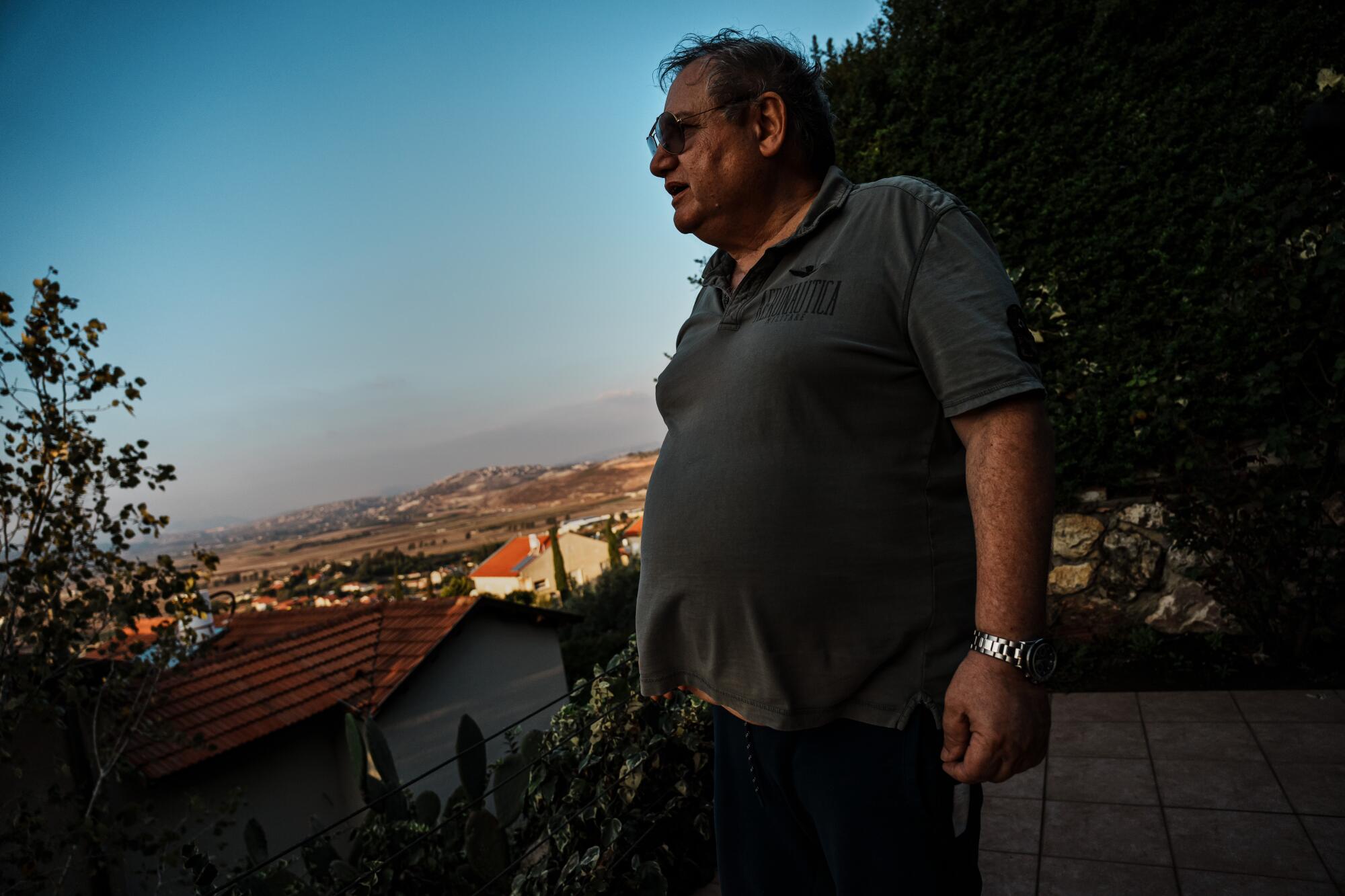 A man wearing glasses and a gray-green shirt looks toward rooftops of homes in a desert landscape 