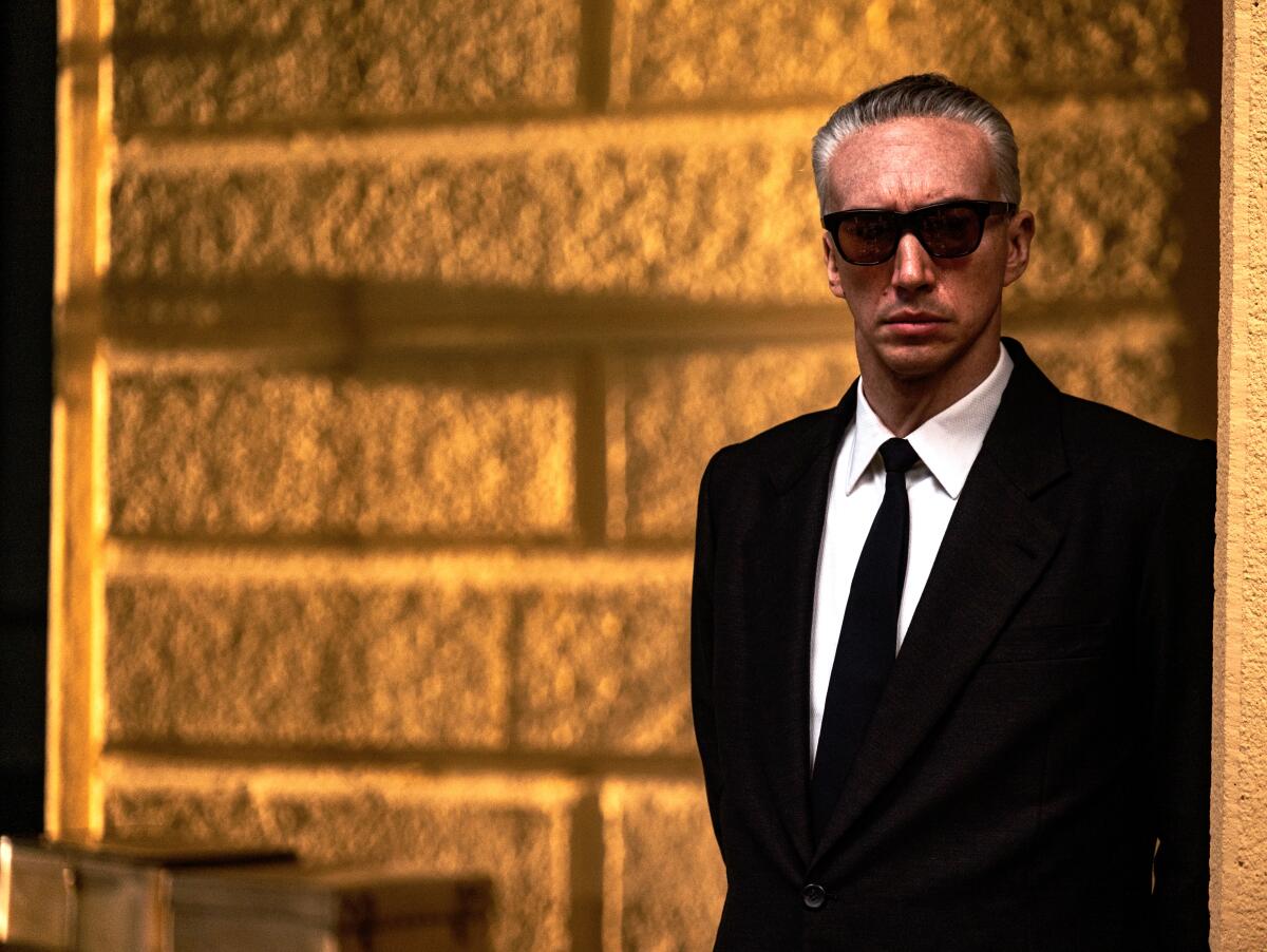 Adam Driver stands in a doorway in sunglasses and business suit on the set of "Ferrari"