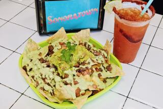 Costilla (steak) nachos on a green tray next to a red michelada and napkin holder that reads "SONORATOWN" at the cantina.