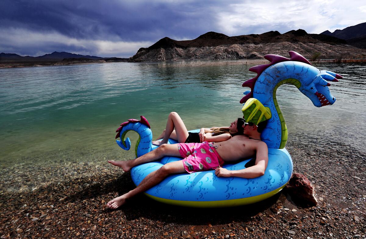 Two people recline on a dragon inflatable floatie next to a body of water
