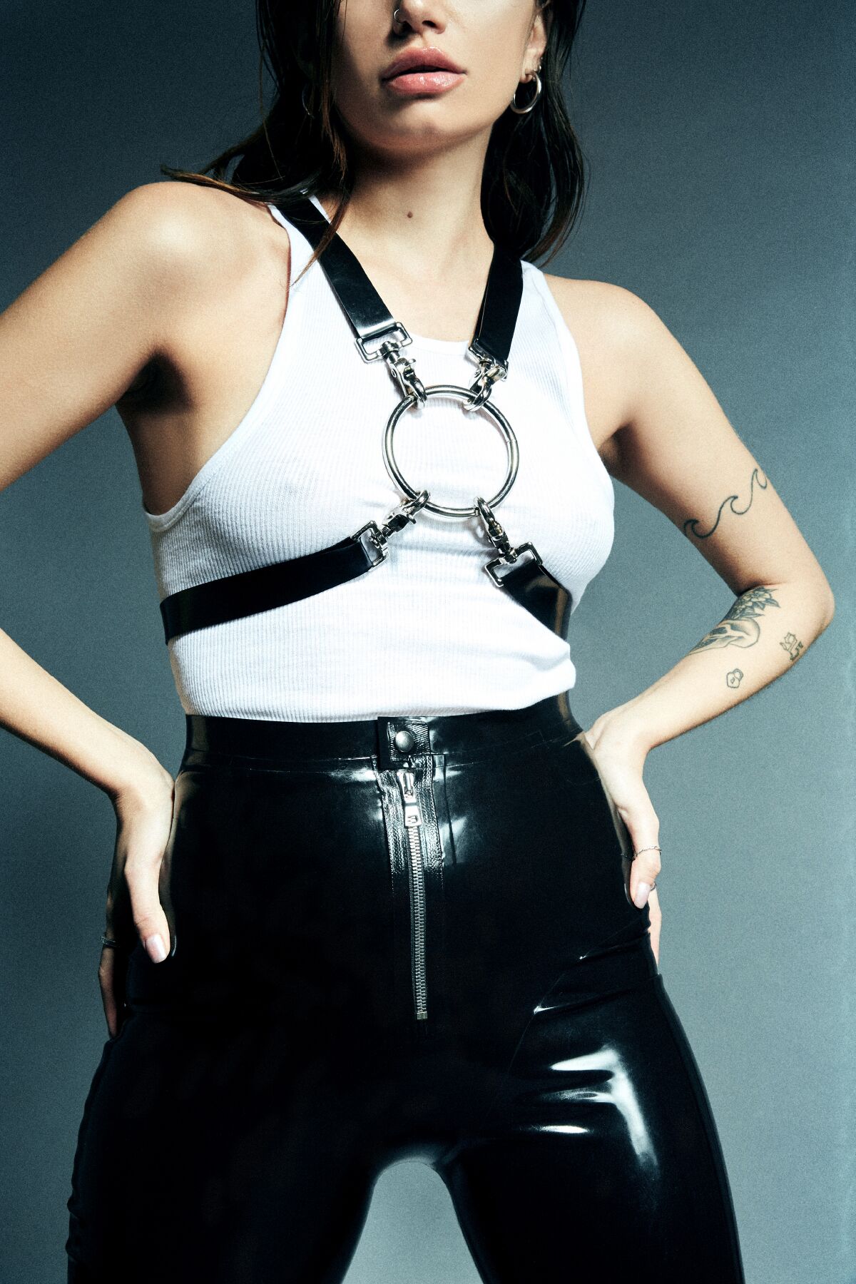 A model wears black pants with a white tank top and a black harness with a large silver ring in the middle.