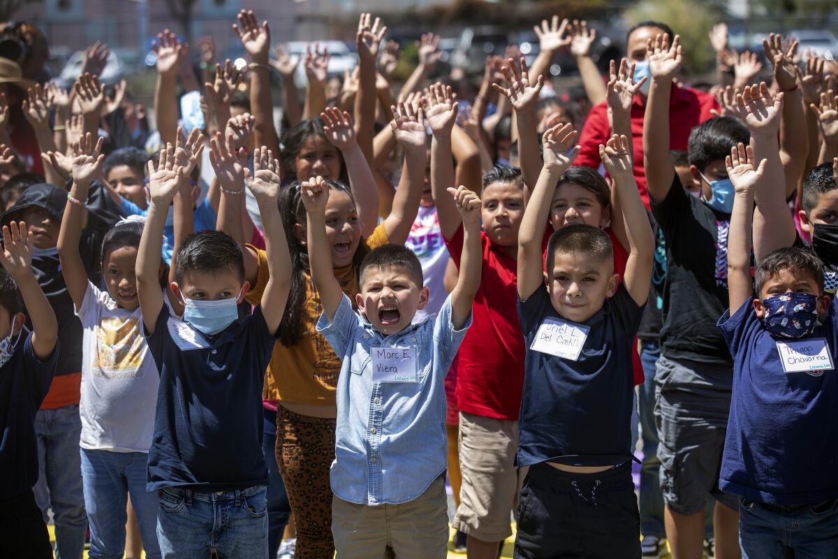 Children, some in masks and some without, raise their hands and shout.