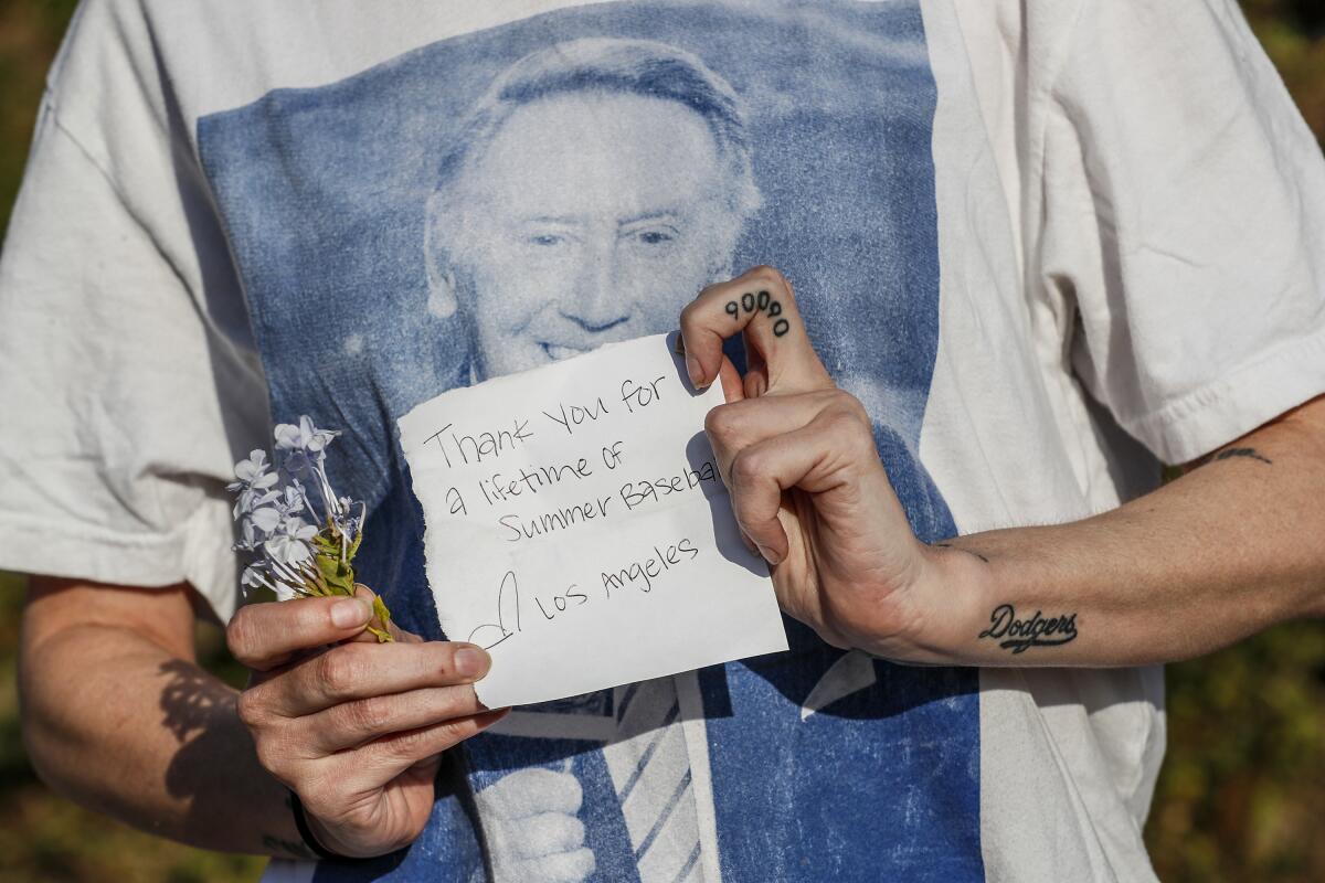 A Dodgers fan wearing a Vin Scully T-shirt holds a note reading, "Thank you for a lifetime of summer baseball" 