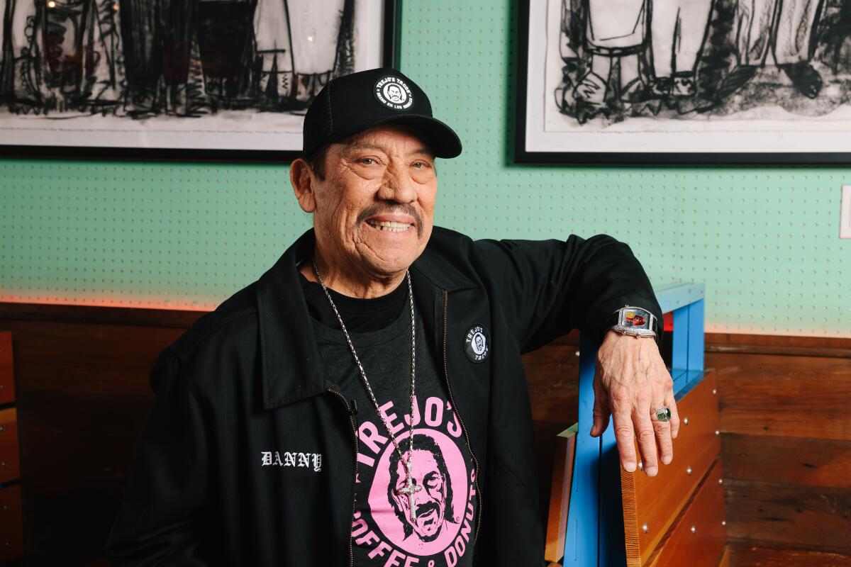 Danny Trejo in a black hat, black shirt with a pink logo and a black jacket sitting inside a restaurant