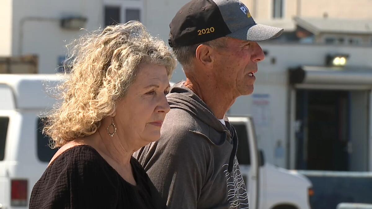 A woman with curly blond hair and a man in a billed cap walk outdoors, their faces grim.