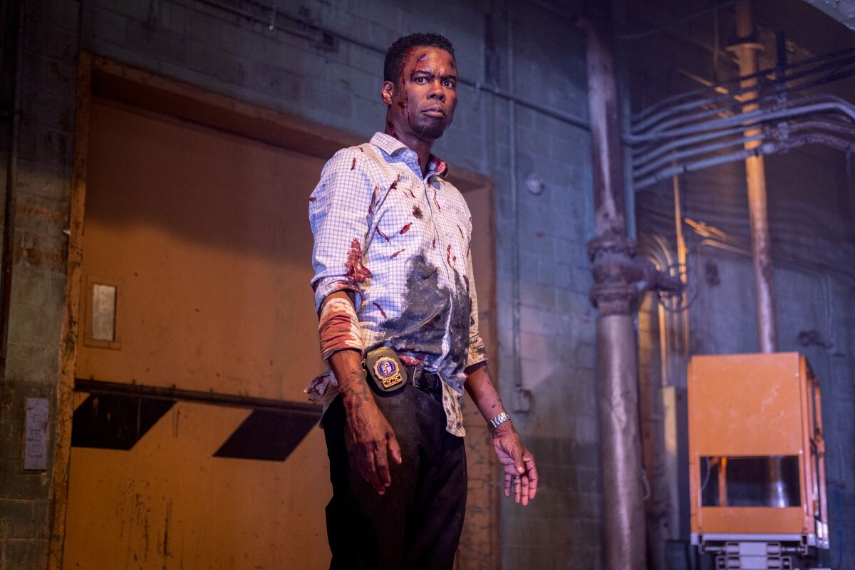 With a policeman's badge at his waist, a battered Chris Rock wears a button-down shirt and dark pants stained with blood.