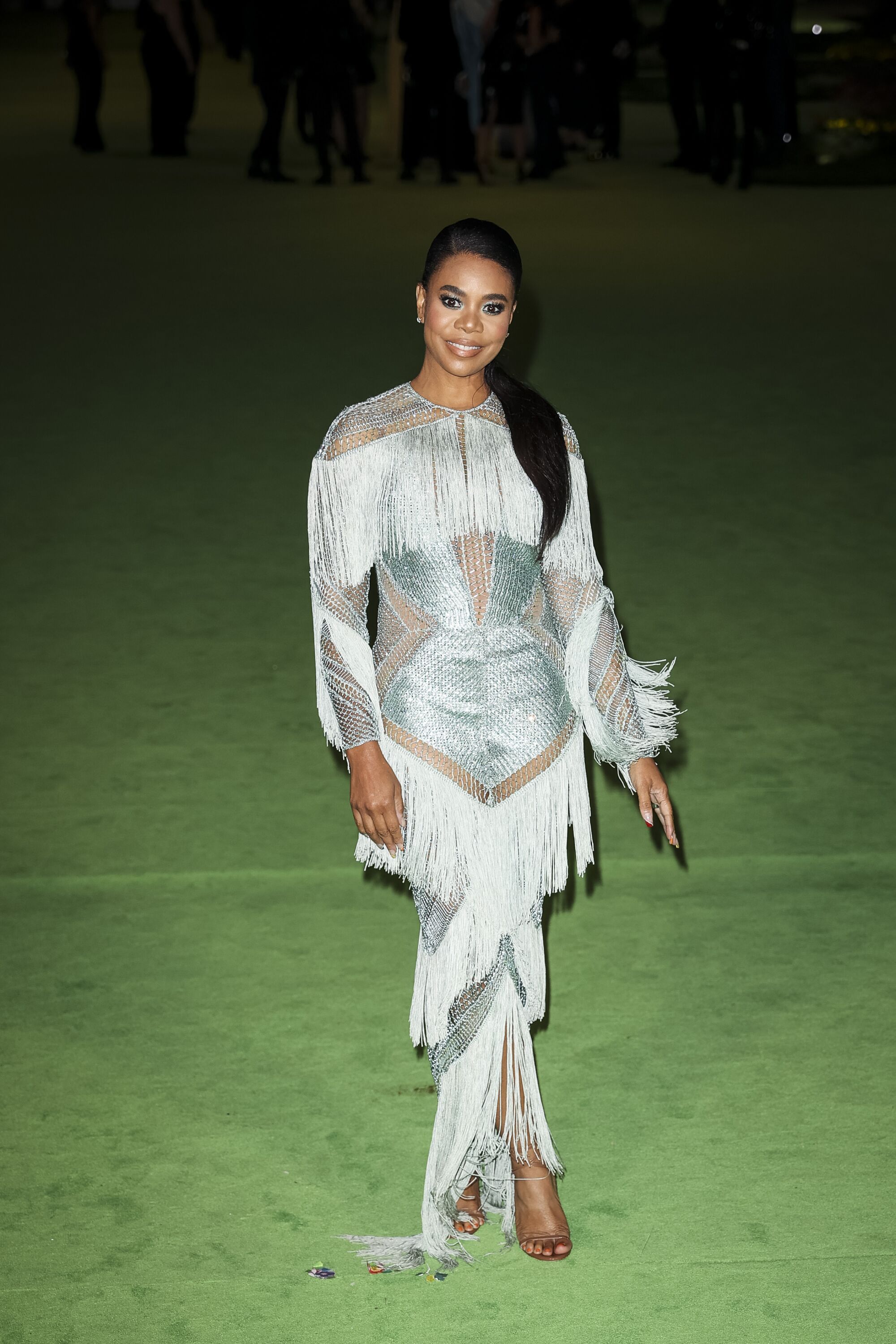 A woman in a silver, fringe dress posing on a green carpet