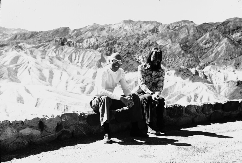 Two men sit on a low wall overlooking desert ridges. 