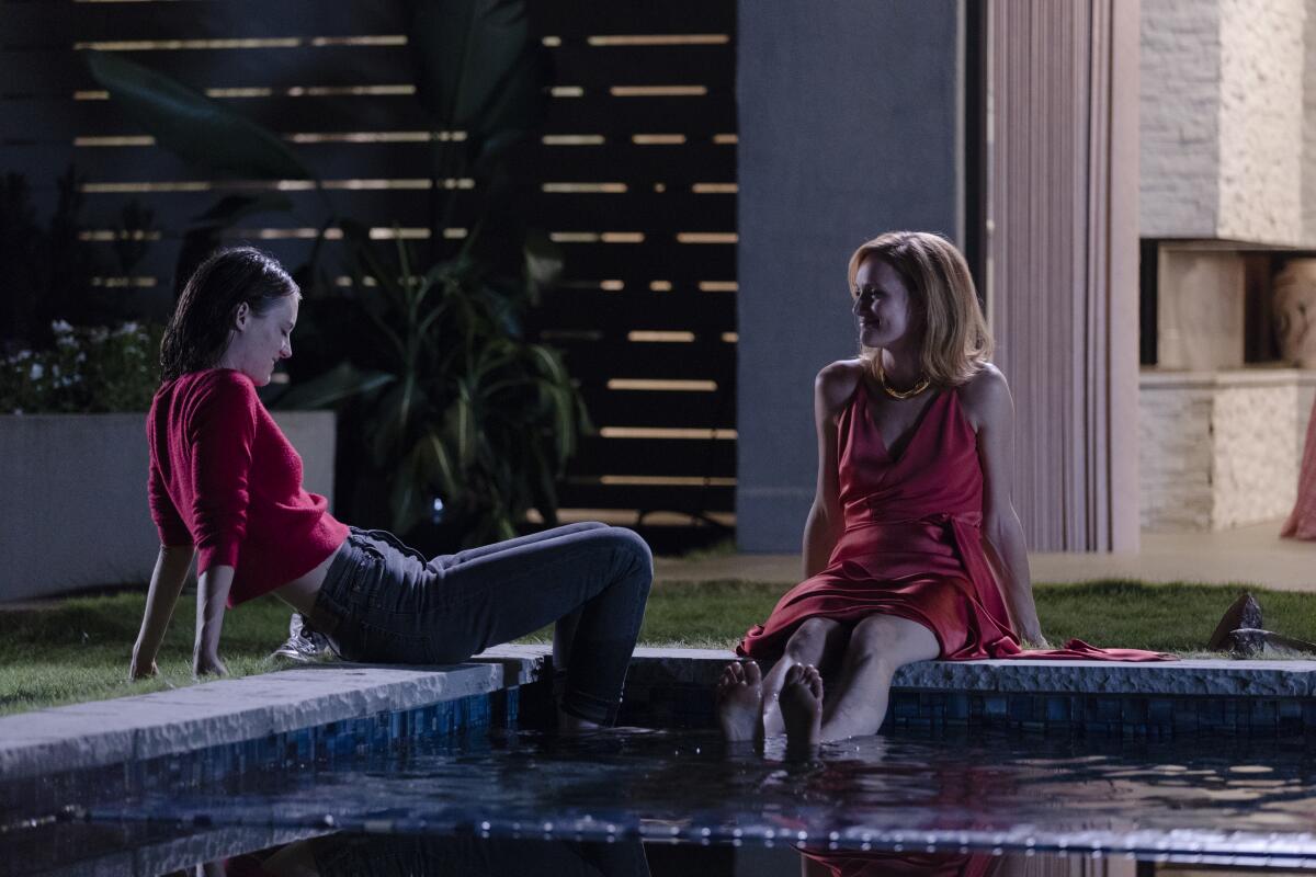 Two women, both in red, sit at the edge of a pool at night.
