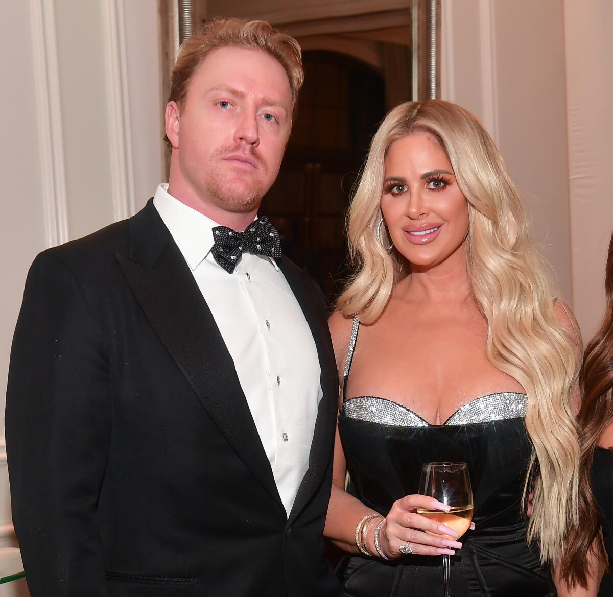 A man in a tuxedo and a woman with long blond hair in a low-cut black dress attend a black-tie gala.