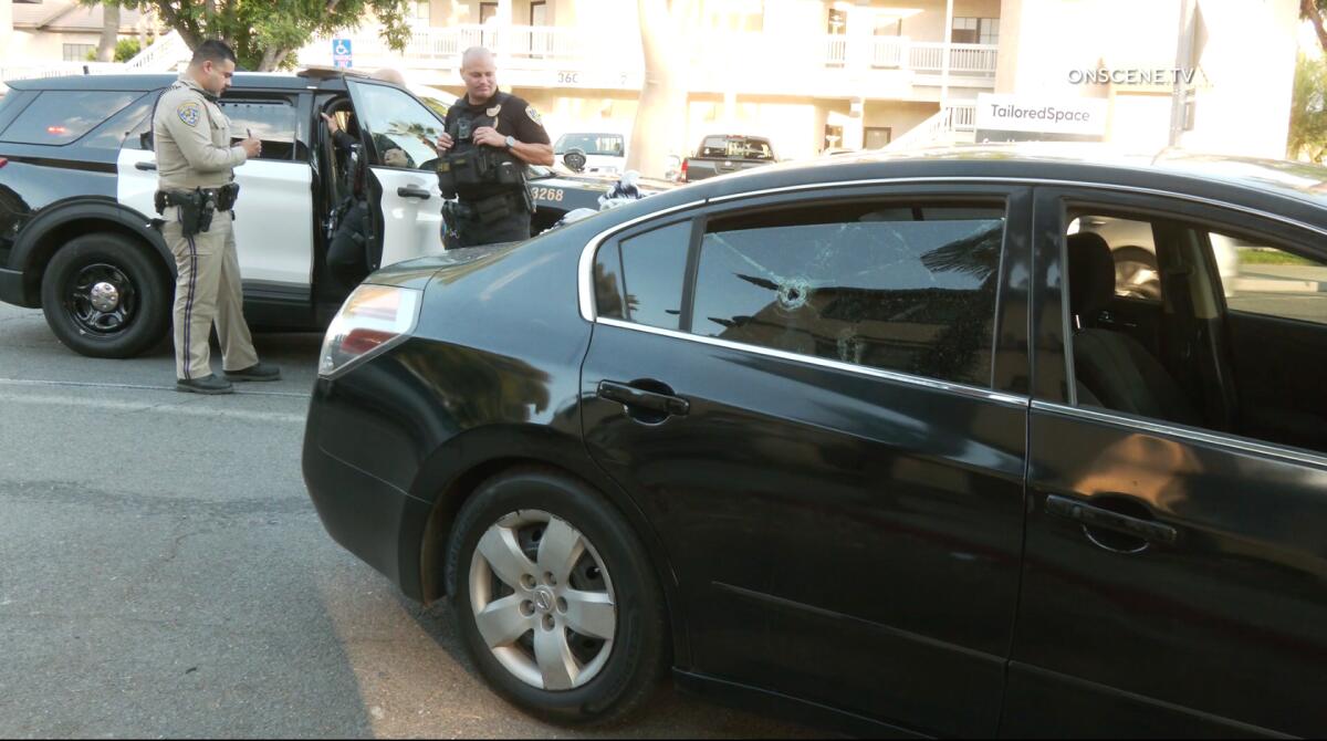 A black car is parked near a law enforcement vehicle on a street with law enforcement standing nearby.