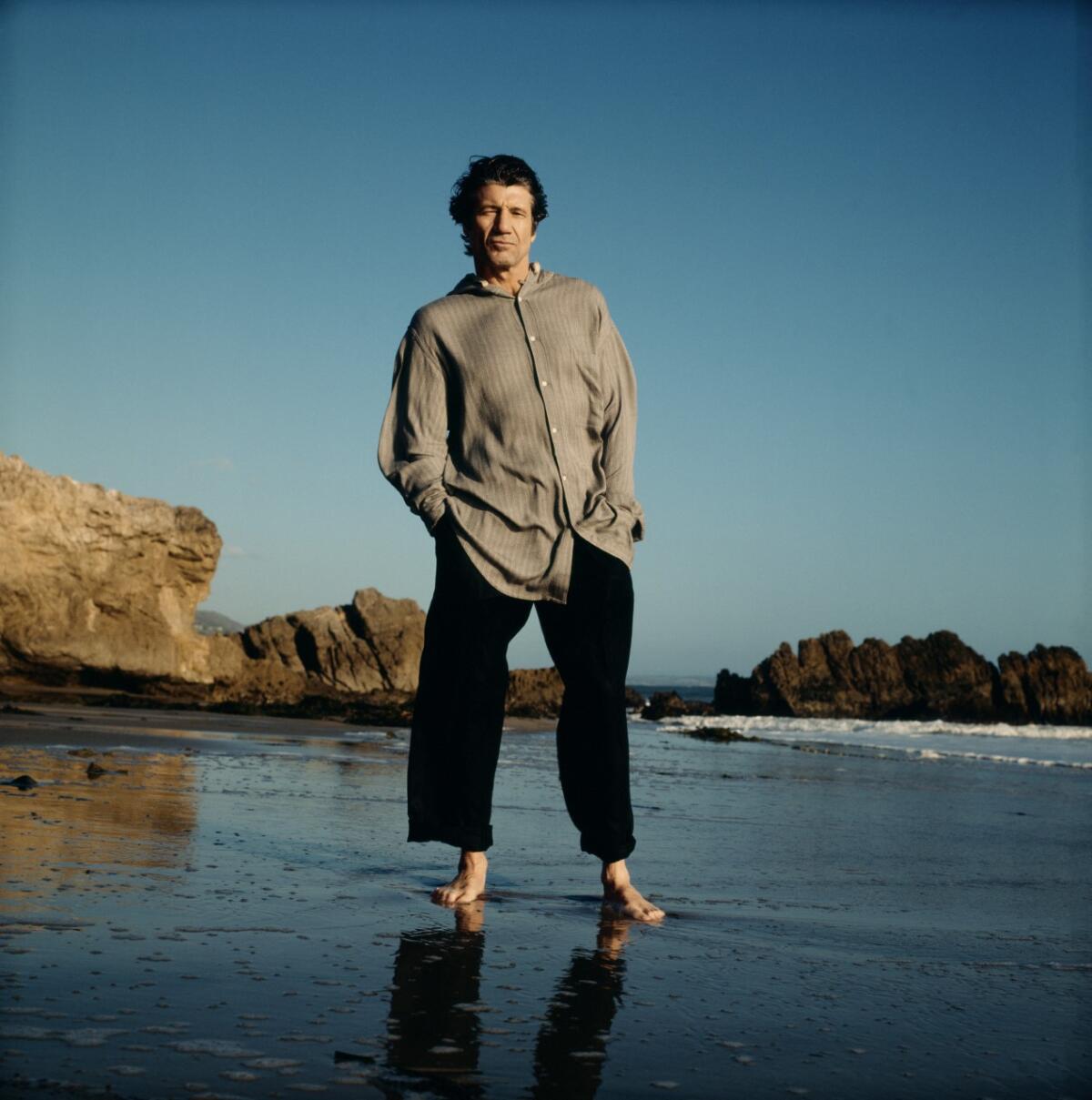 A man in loose clothing and bare feet stands on a beach with rocks behind him