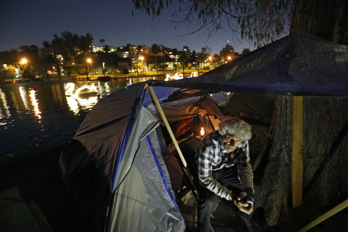 A man sits in the entrance of a tent at the edge of a lake at night as city lights reflect in the water
