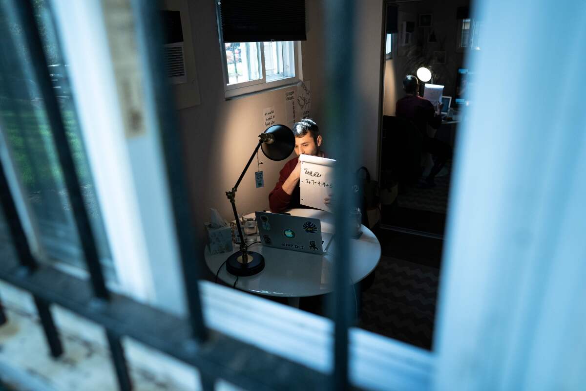 A teacher at his computer is glimpsed through the security-barred windows of his apartment.  