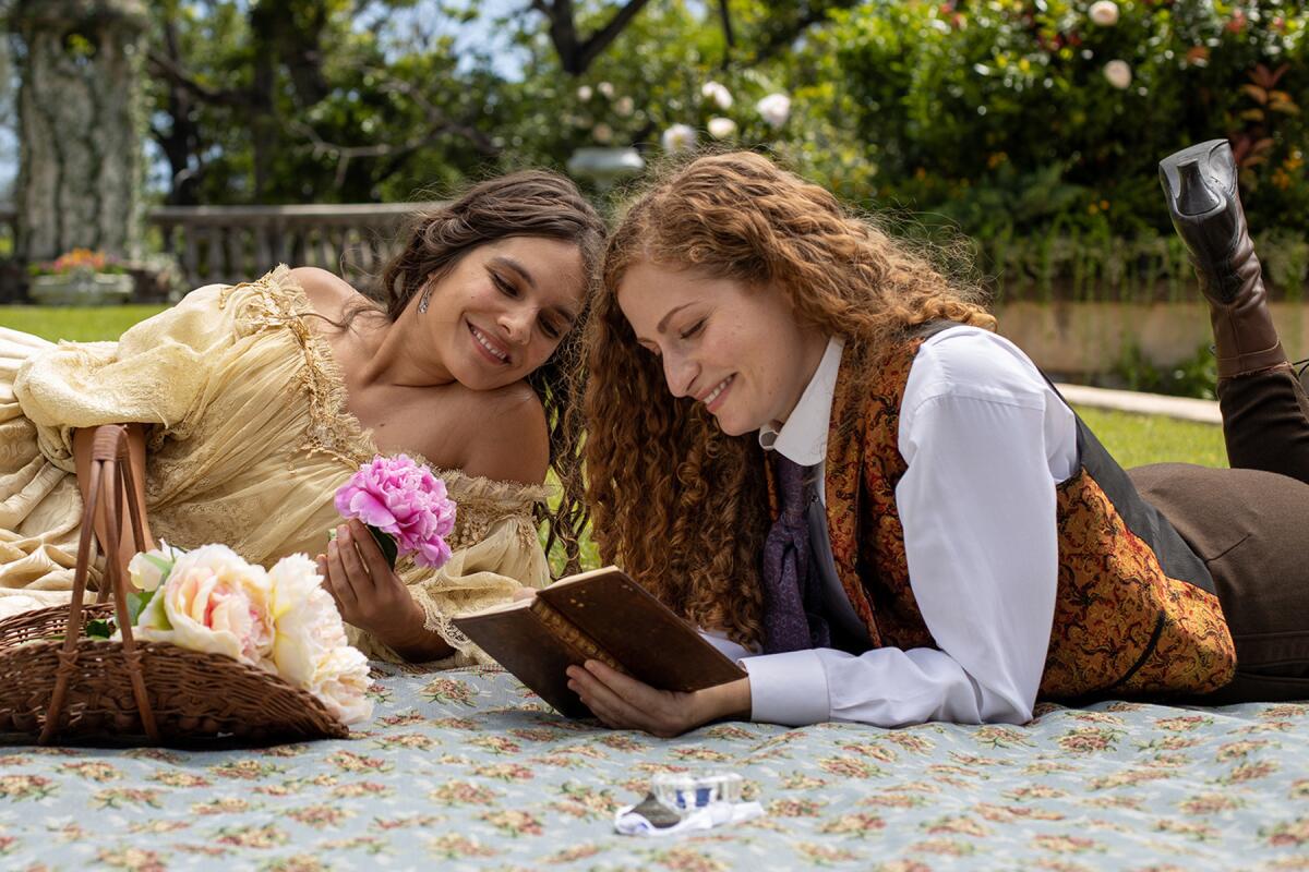 Two women look at an open book while lying on the ground on a blanket.