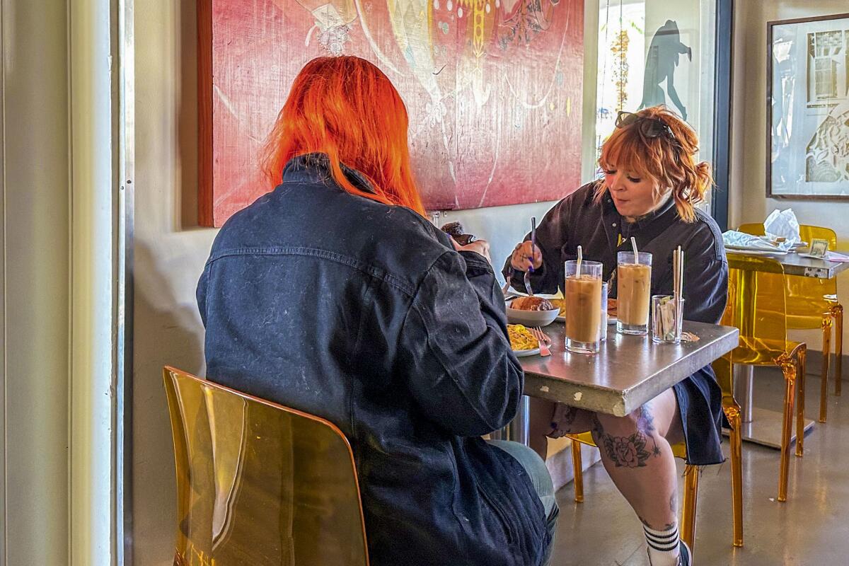 Two people at a restaurant table with colorful paintings on the wall above them.