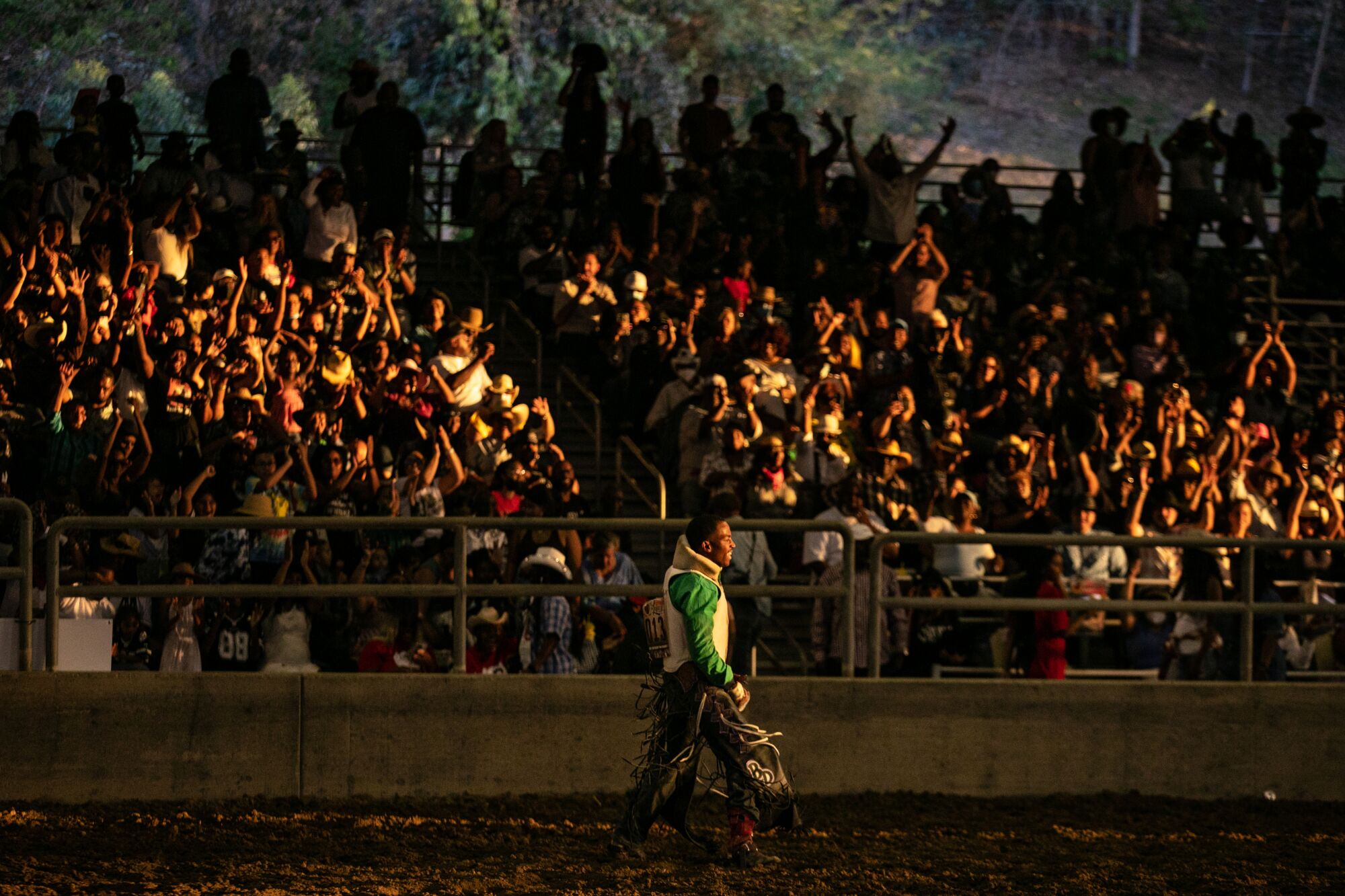 A crowd cheering from the stands as a man carrying a saddle walks through a rodeo arena