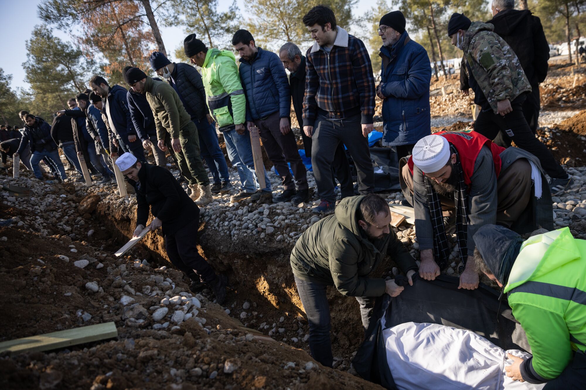 A small crowd standing at the edge of a trench in the dirt as men place a body covered in white fabric inside