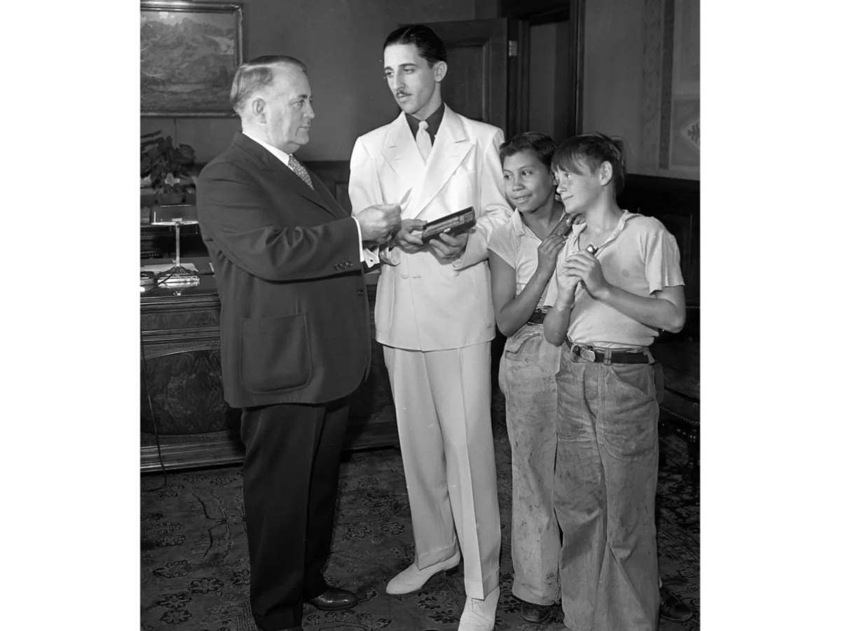 A serious older man turns to a young man and two boys holding harmonicas