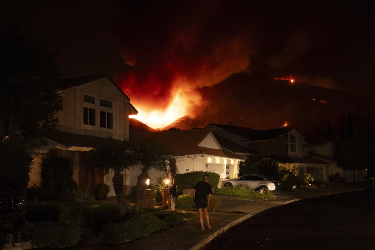 A man in a front yard watches flames on the ridge nearby.