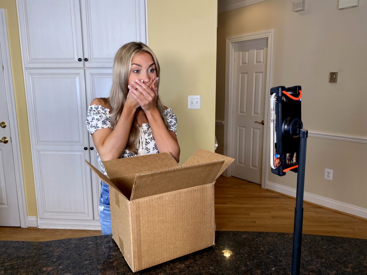A woman with her hands over her mouth in front of an open box and a cellphone on a stand