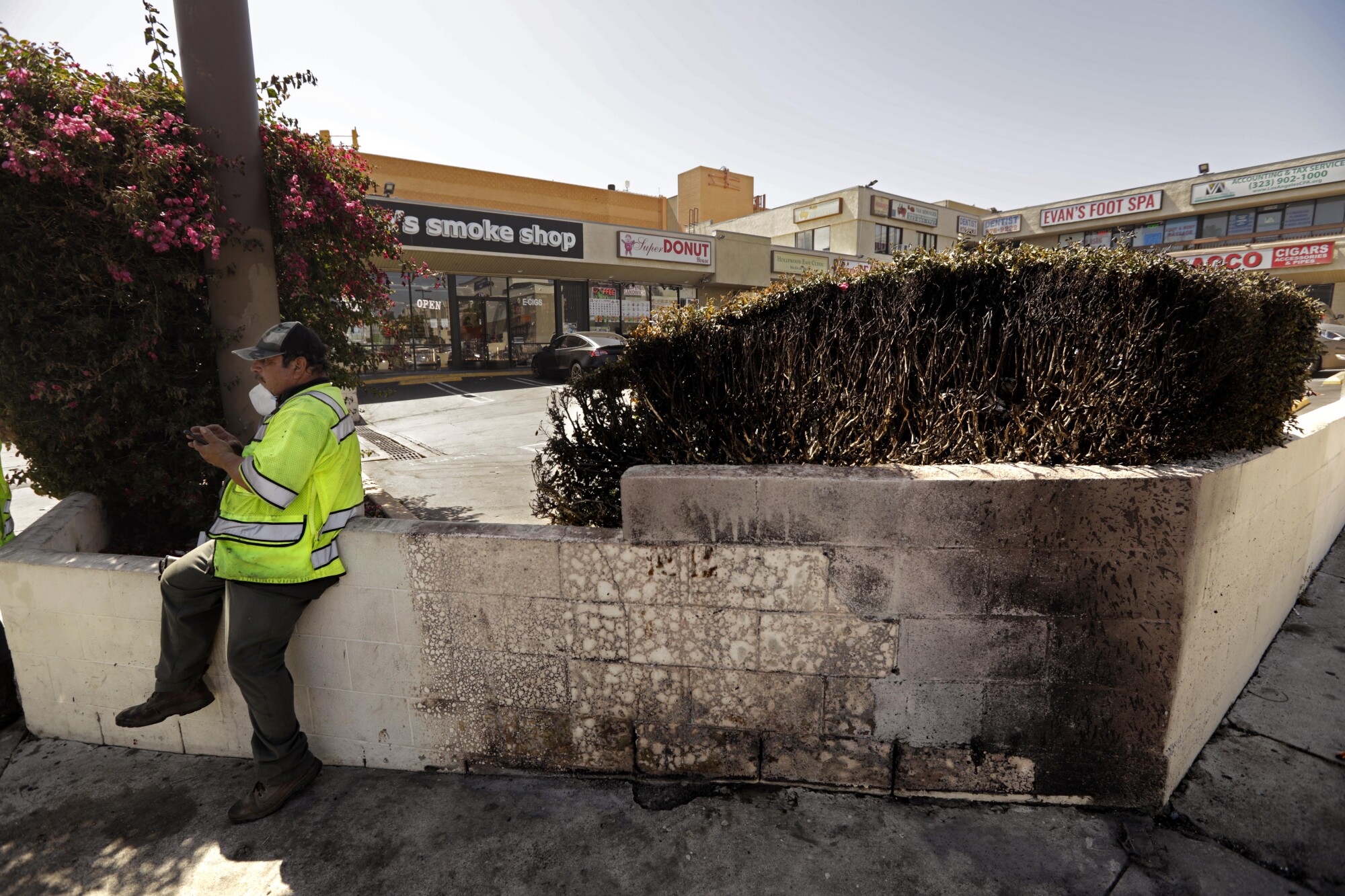 A sanitation worker rests next to a wall and bush that were charred by a fire 