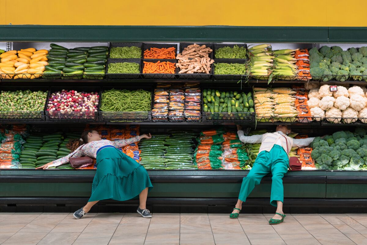 Two women sprawl across vegetables in a supermarket produce aisle.