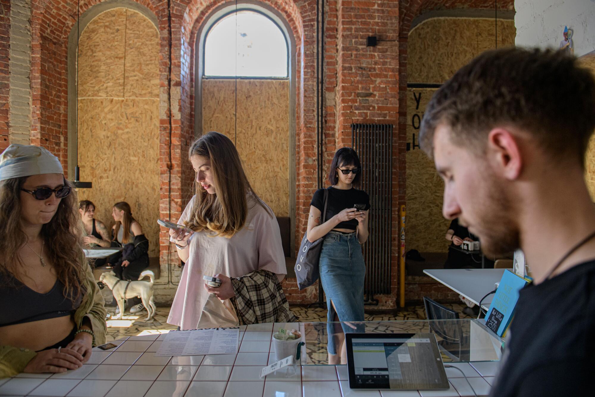 A girl looks at her phone near other people in a coffeehouse with brick walls and boarded-up windows