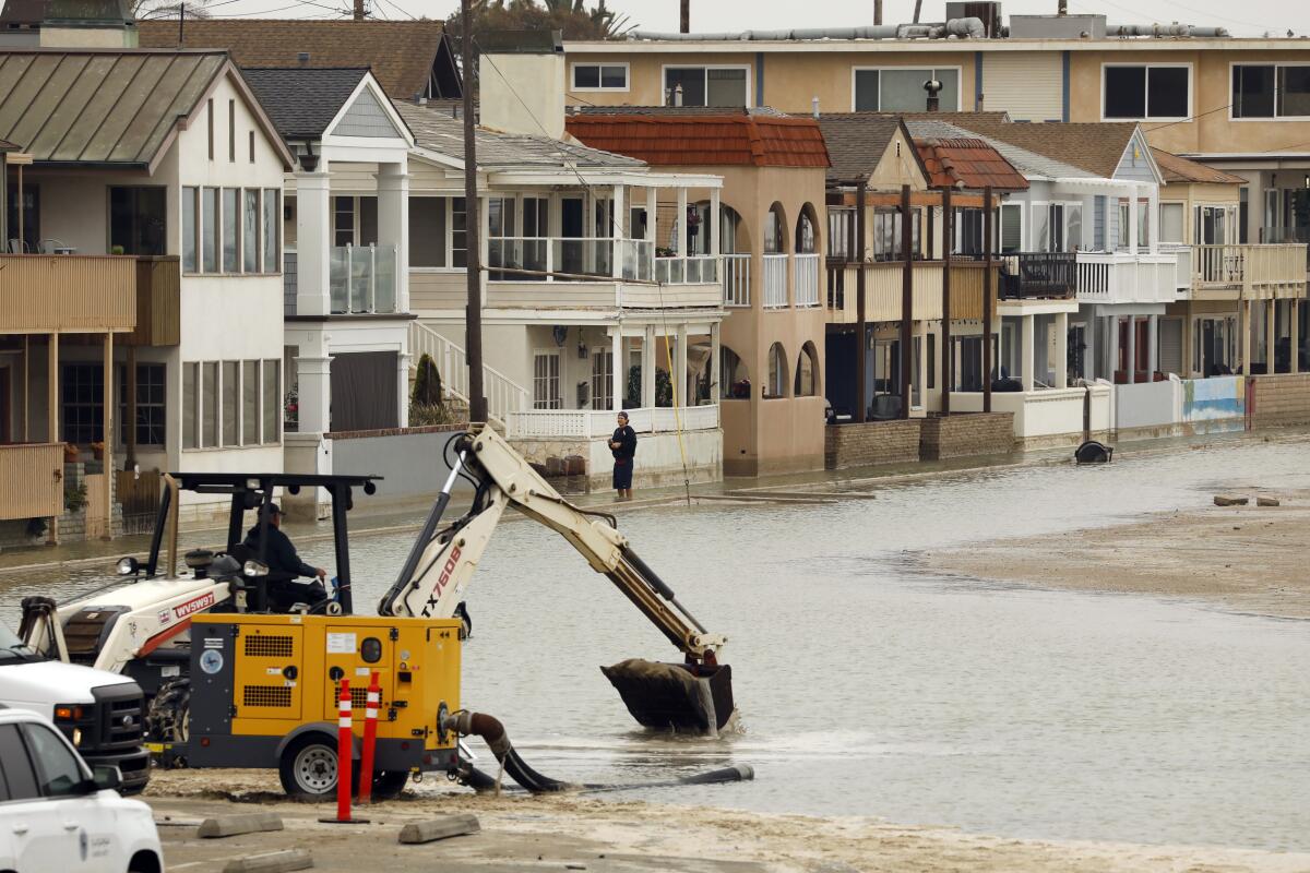 A backhoe scoops waterlogged sand from a beach next to a row of homes