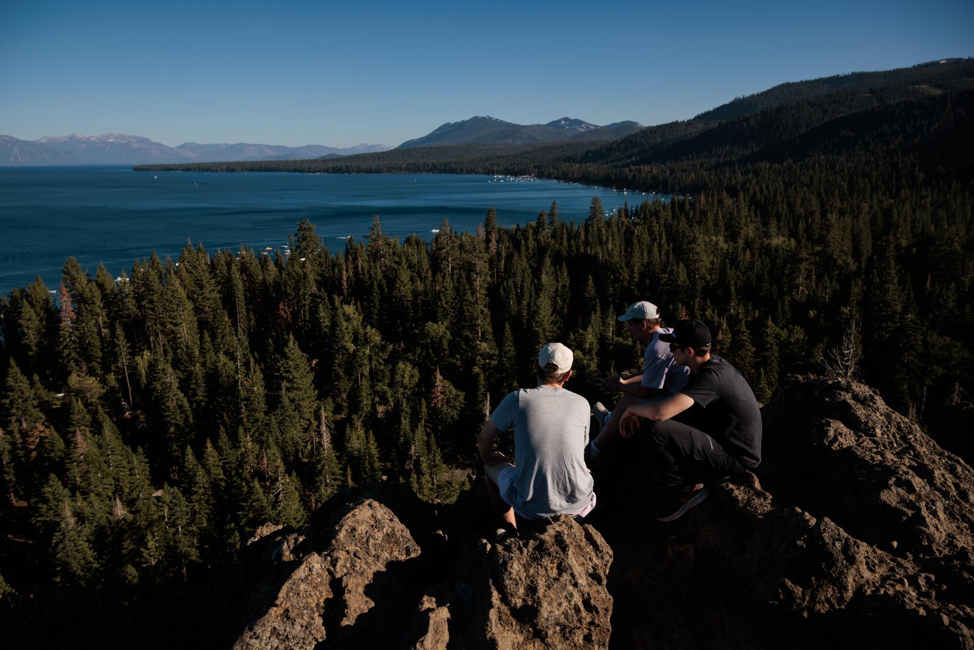 Three people perch on a rock and gaze out over conifers toward an alpine lake.