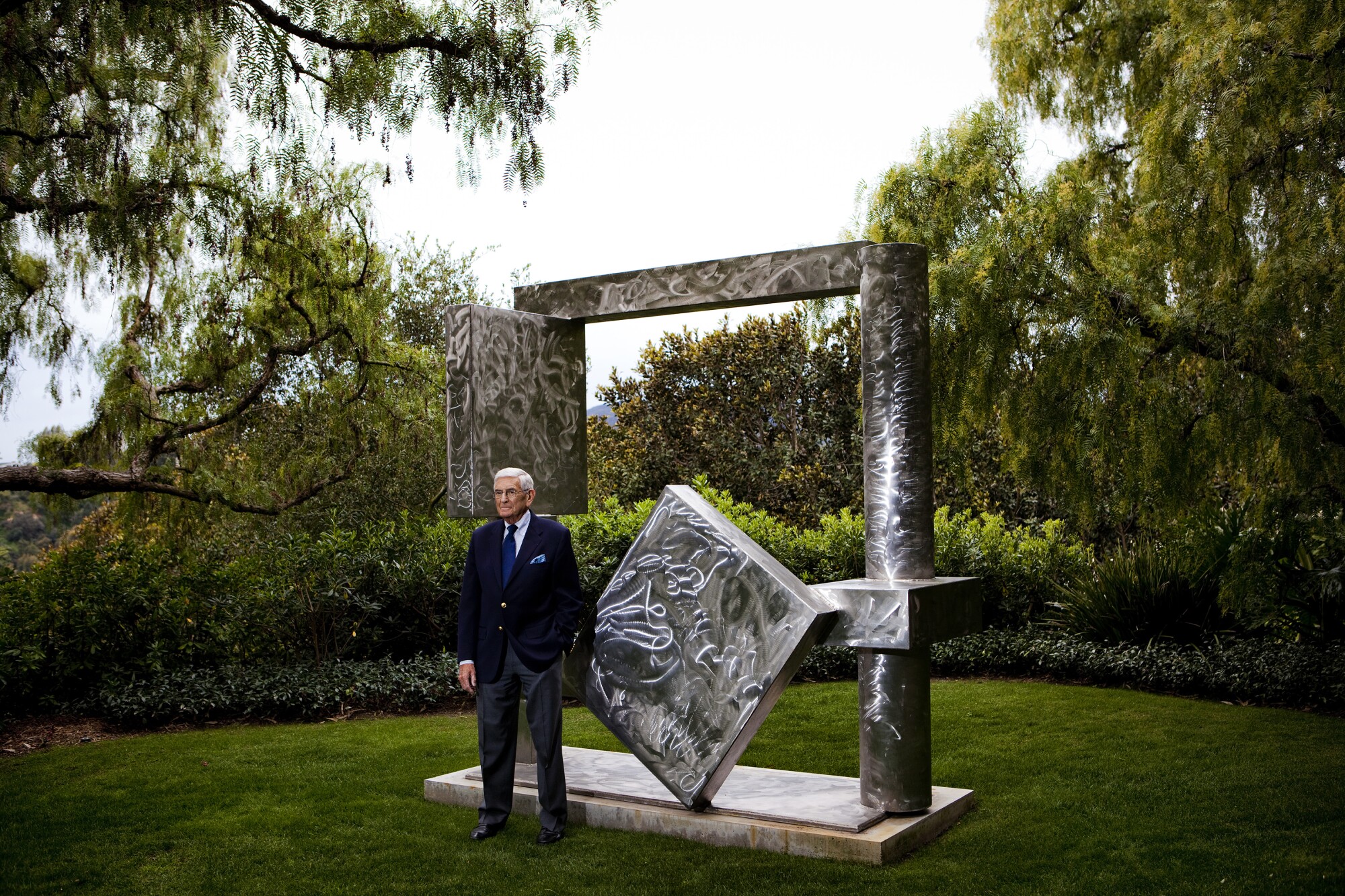 Eli Broad stands in front of a metal sculpture against a backdrop of shrubs and trees.
