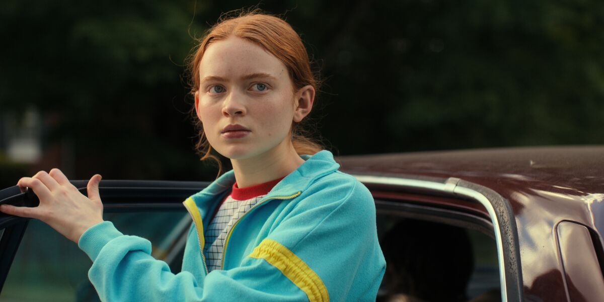 A girl with red hair wearing a blue jacket and holding a car door open
