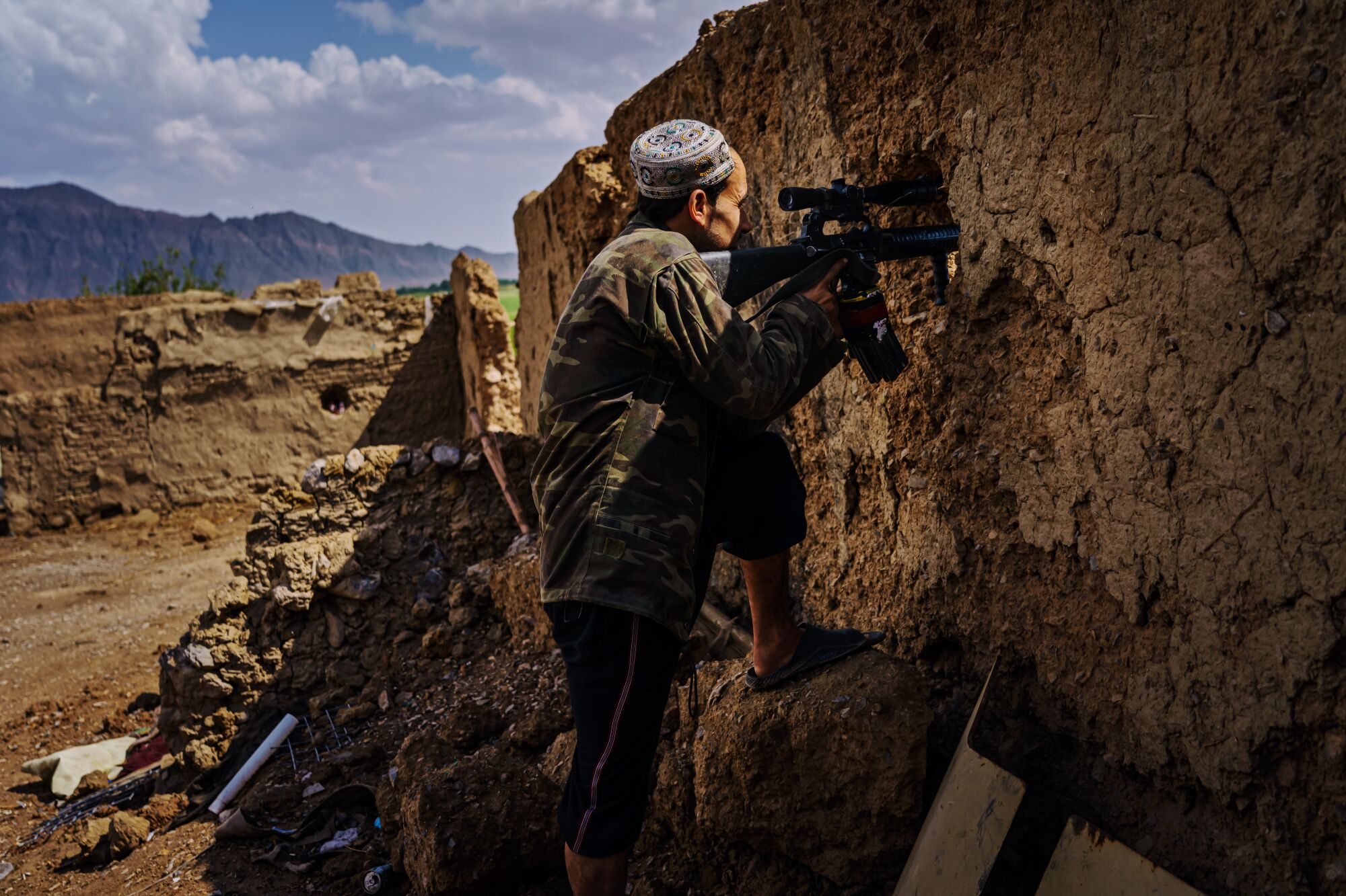 Azimullah Muhamadi holds a weapon and surveys the terrain through a hole in a wall with a sniper scope